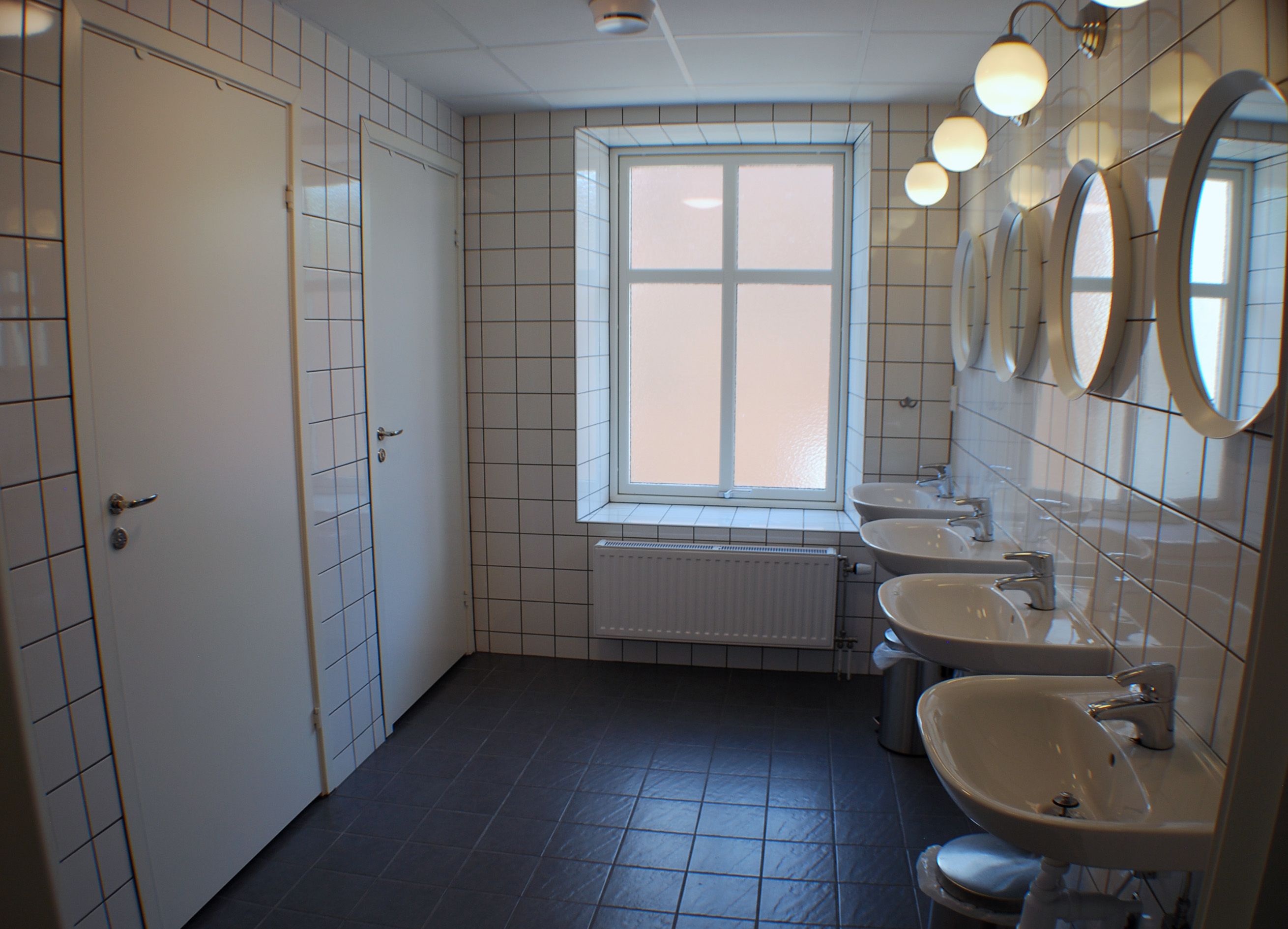 Un baño con paredes de azulejos blancos, una ventana y tres lavamanos.