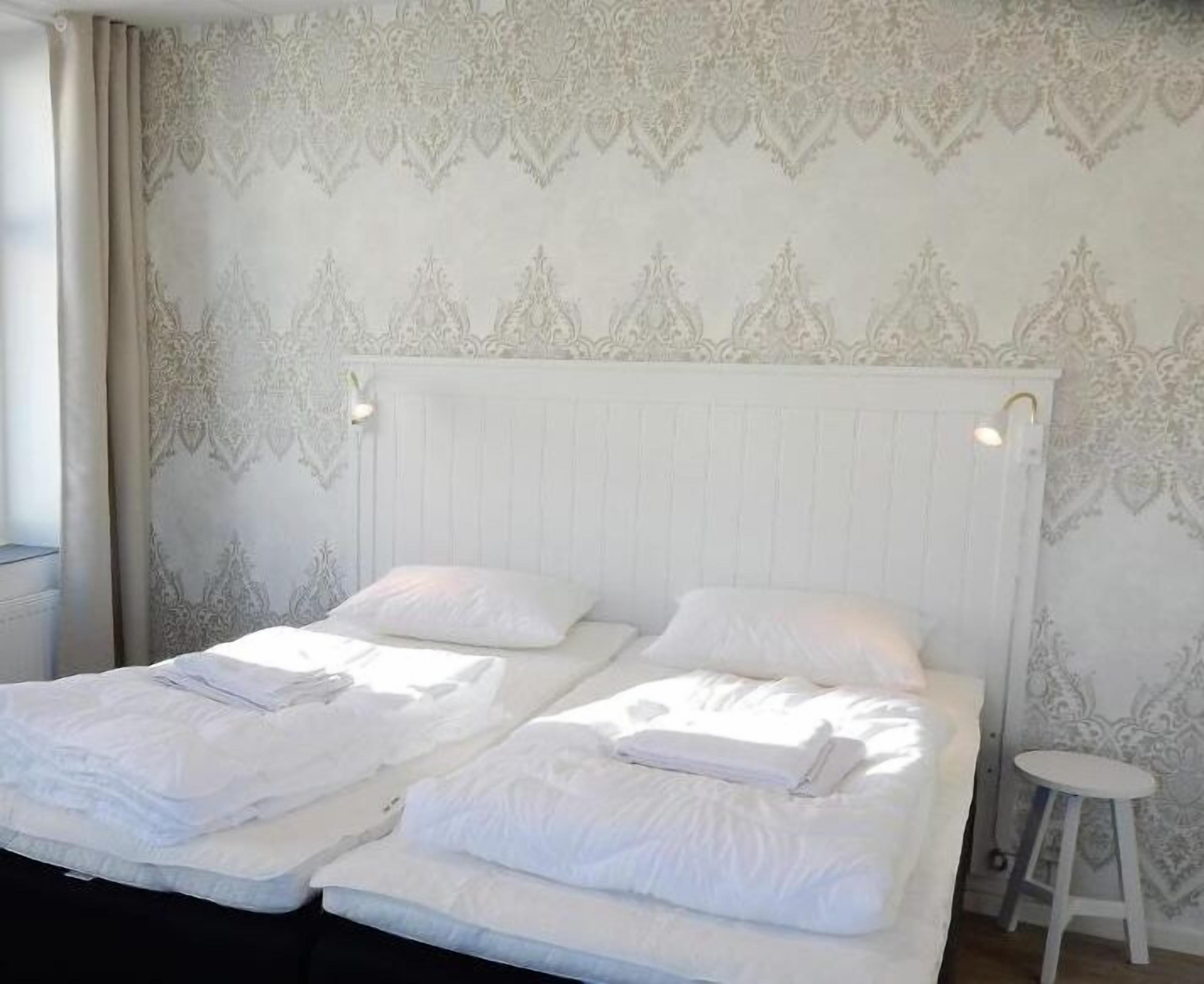 A double bed with white linens and two pillows, a white nightstand, and a white stool.