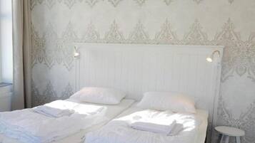 Cama doble con ropa de cama blanca y dos almohadas, mesita de noche blanca y taburete blanco.