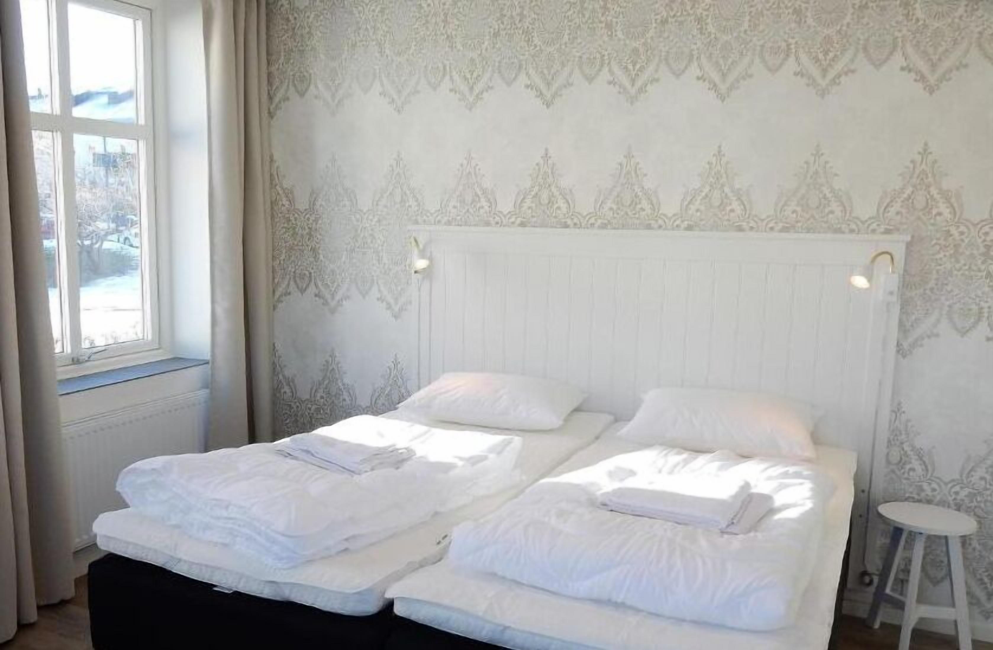Habitación con dos camas, ropa de cama blanca, un radiador, papel tapiz estampado y una ventana con cortinas.