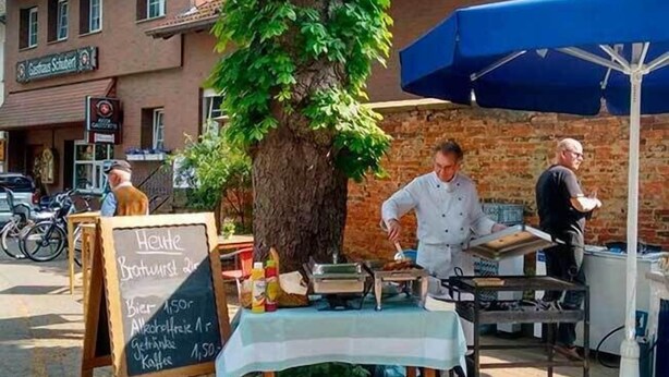A street-side food stall with a chalkboard menu, a chef preparing food, and a brick building in the background. - Hotel Gasthaus Schubert (Garbsen)