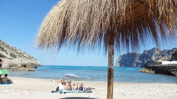 A beach scene with a thatched umbrella, rocky cliffs, and clear blue sea. - Apt. Grande Ciruelo, A/C, community pool, Wifi, near beach 400 m (Cala Sant Vicenç)