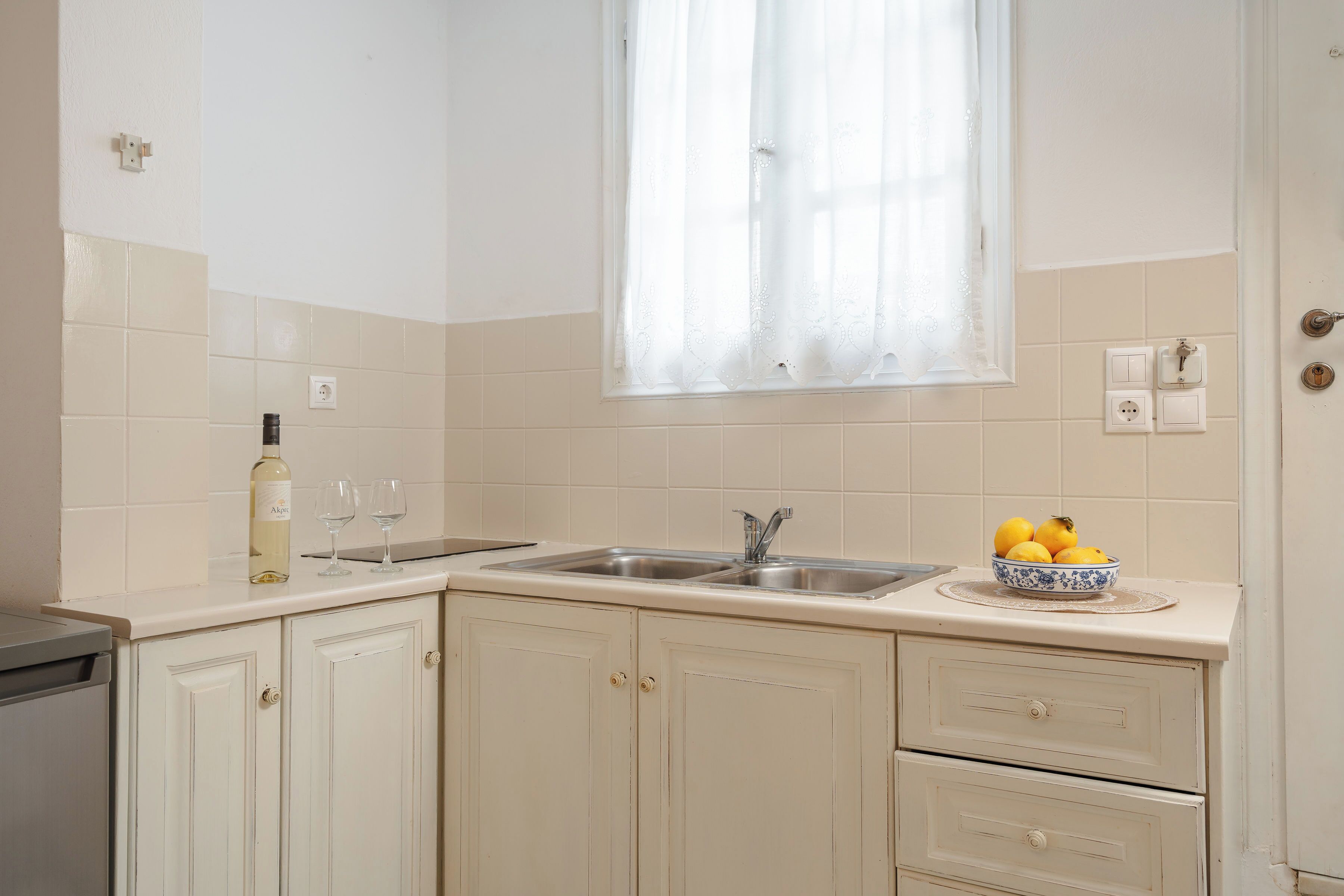 Cuina amb armaris blancs, una bateda doble i una finestra amb cortines de tela fina.