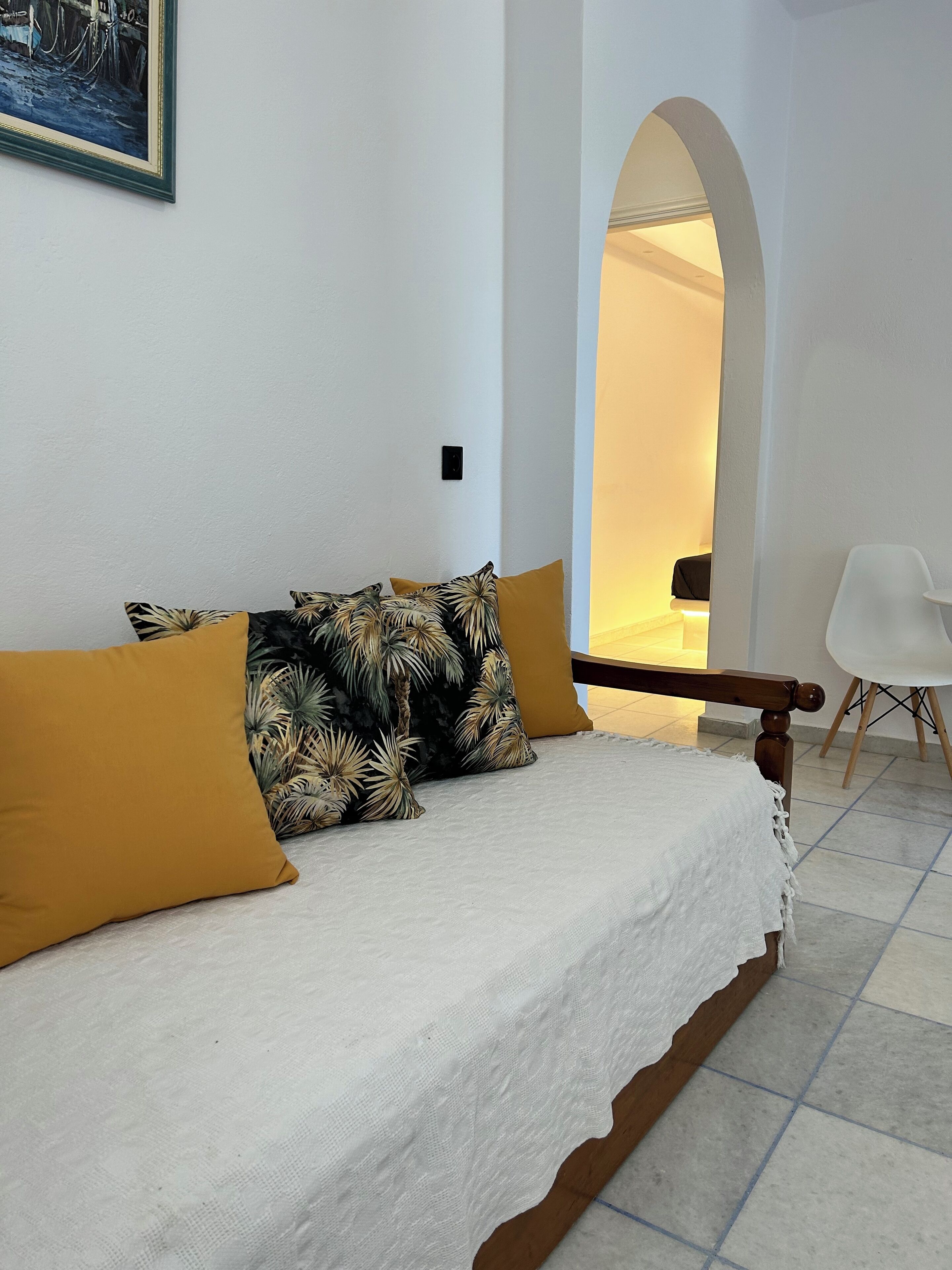 A bed with a white bedspread and two pillows, one yellow and one with a palm leaf pattern, in a room with white walls and a tiled floor.