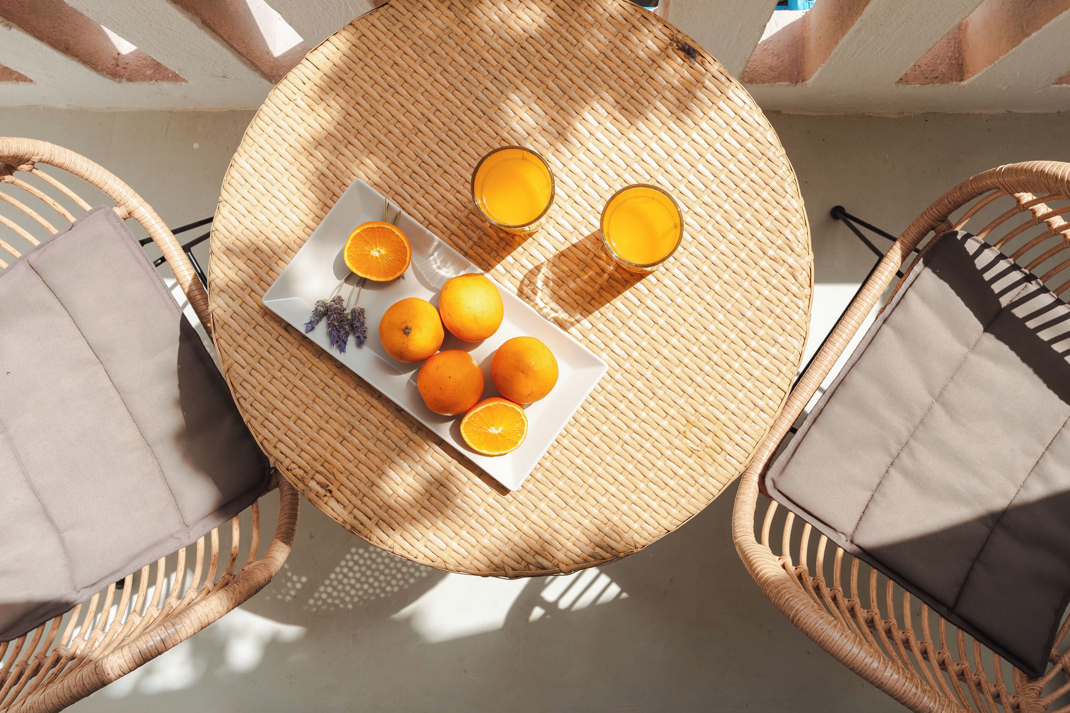 Una mesa redonda de mimbre con un plato de naranjas y dos vasos de jugo de naranja, rodeada de sillas de mimbre con cojines.