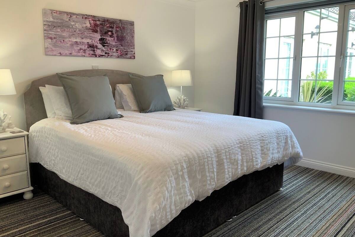 A neatly made bed with a white duvet and grey pillows, a bedside table with a lamp, and a window with curtains.