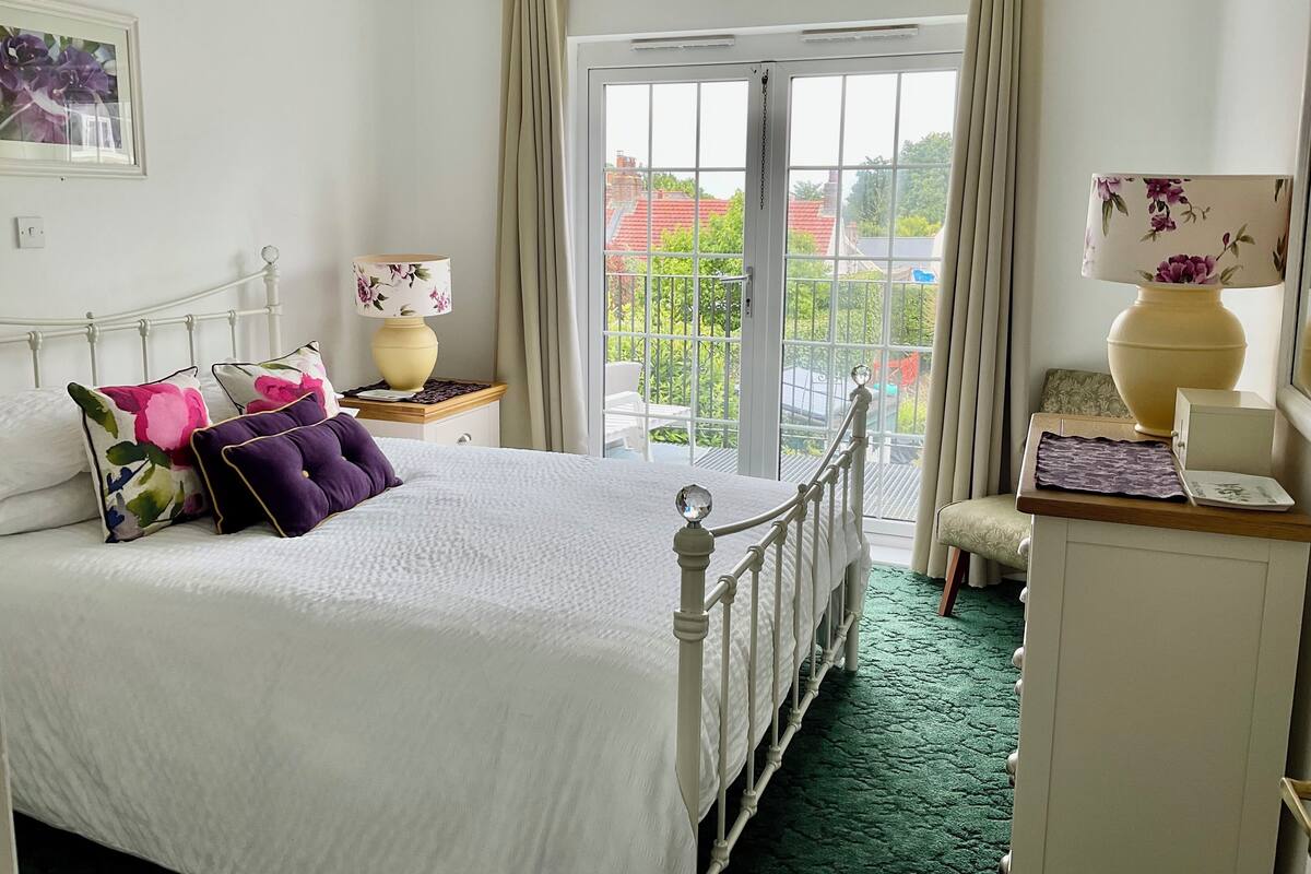 A bedroom with a bed, a nightstand, a lamp, a chair, and a view of the outside.