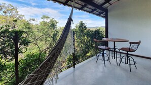 Una hamaca en un balcón con vistas a árboles y colinas. Hay una mesa y dos sillas en el balcón.