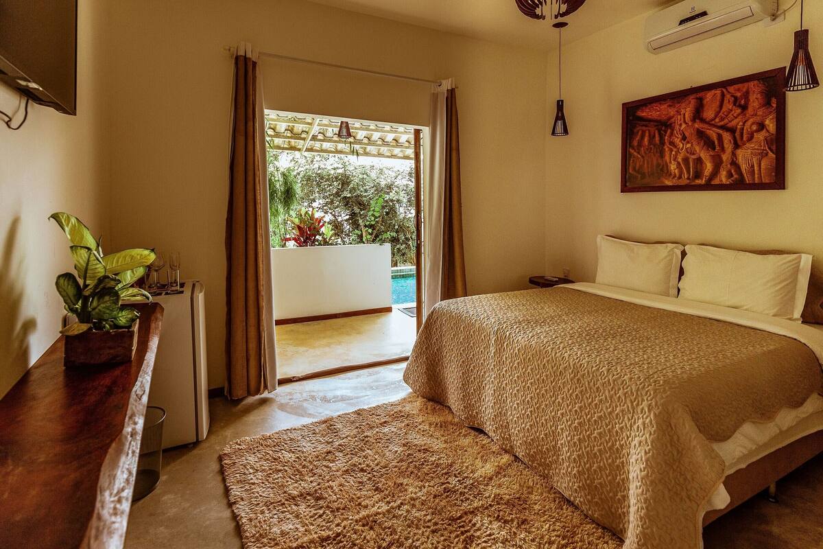 A bedroom with a bed, a wooden nightstand, a potted plant, and a view of the outdoors.