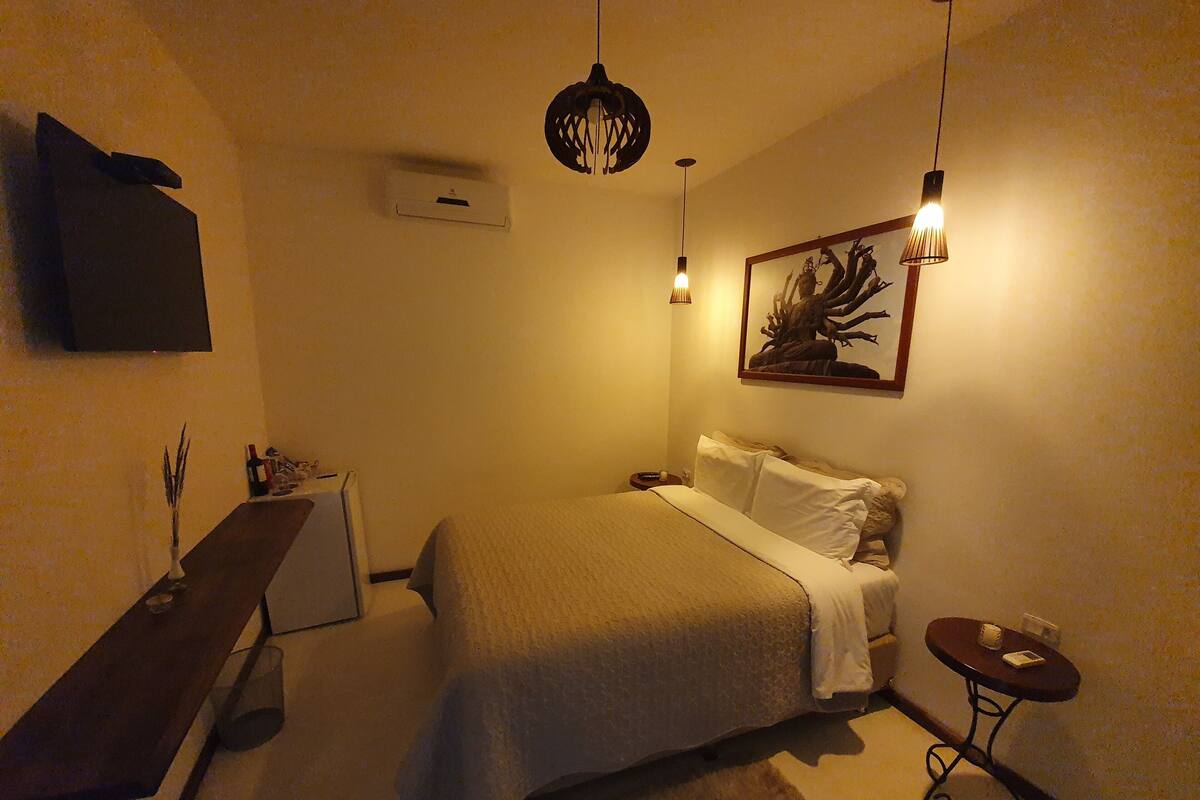 A bedroom with a bed, bedside table, lamp, artwork, and a flat-screen TV mounted on the wall.