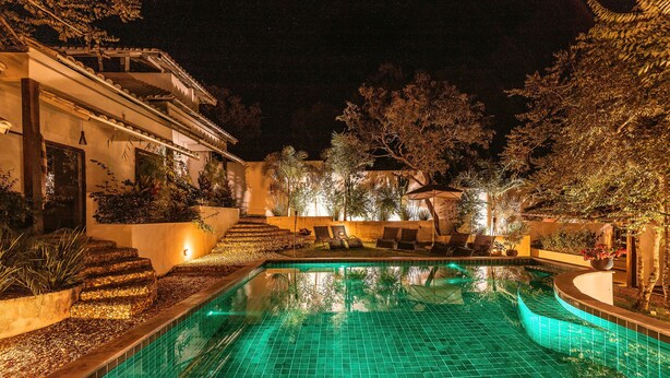 Vista nocturna de una piscina con suelo de baldosas, rodeada de un jardín ajardinado y un edificio con escaleras que conducen a una puerta.