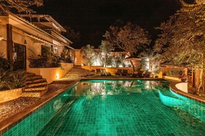 Vista nocturna de una piscina con suelo de baldosas, rodeada de un jardín ajardinado y un edificio con escaleras que conducen a una puerta.