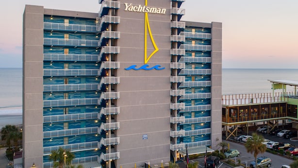 A multi-story hotel with a wave-themed facade, named "Yachtsman," featuring balconies and a view of the ocean.