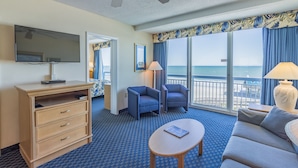 A hotel room with a large window, a ceiling fan, a flat-screen TV, a wooden dresser, two armchairs, a sofa, and a small table.