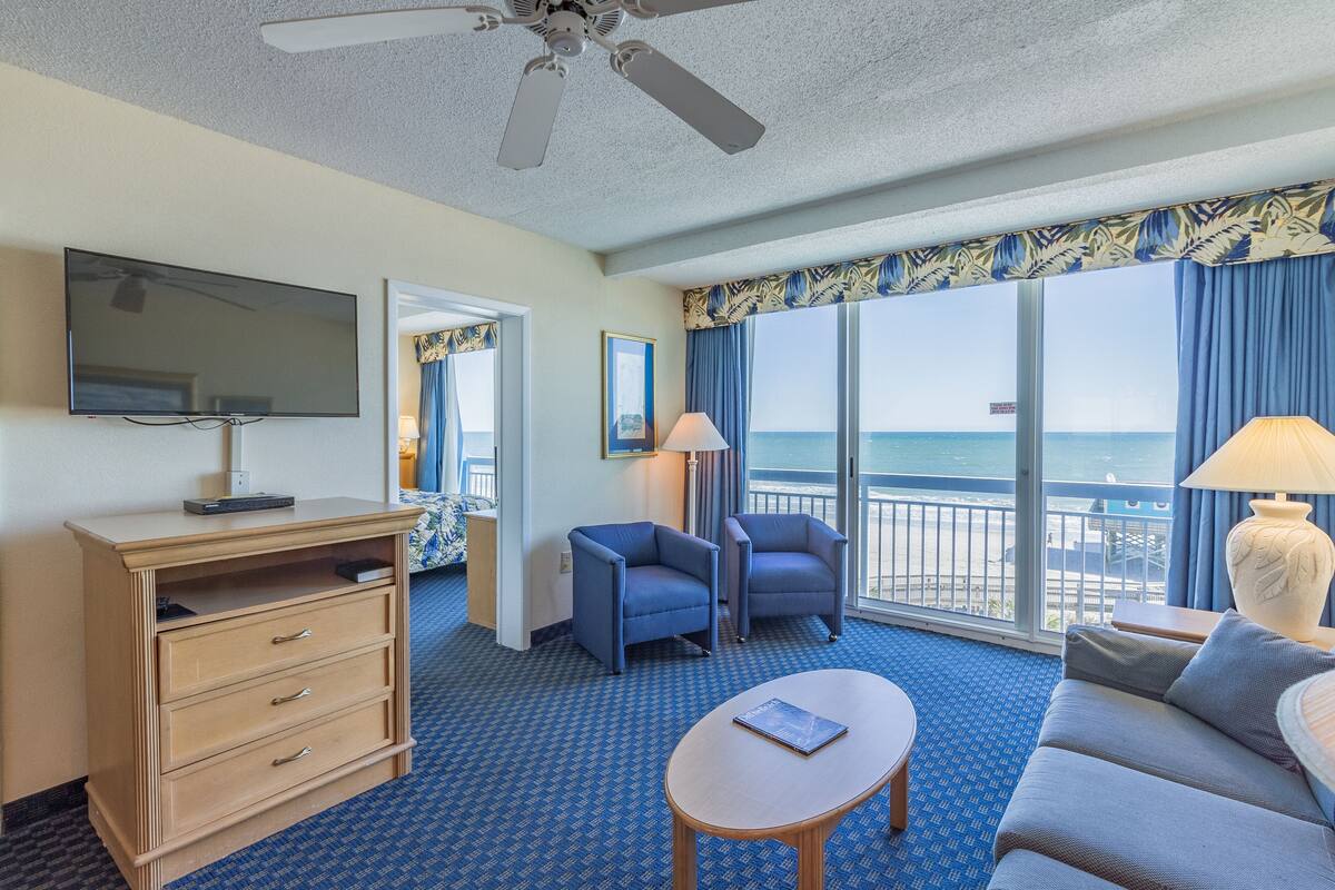 A hotel room with a large window, a ceiling fan, a flat-screen TV, a wooden dresser, two armchairs, a sofa, and a small table.