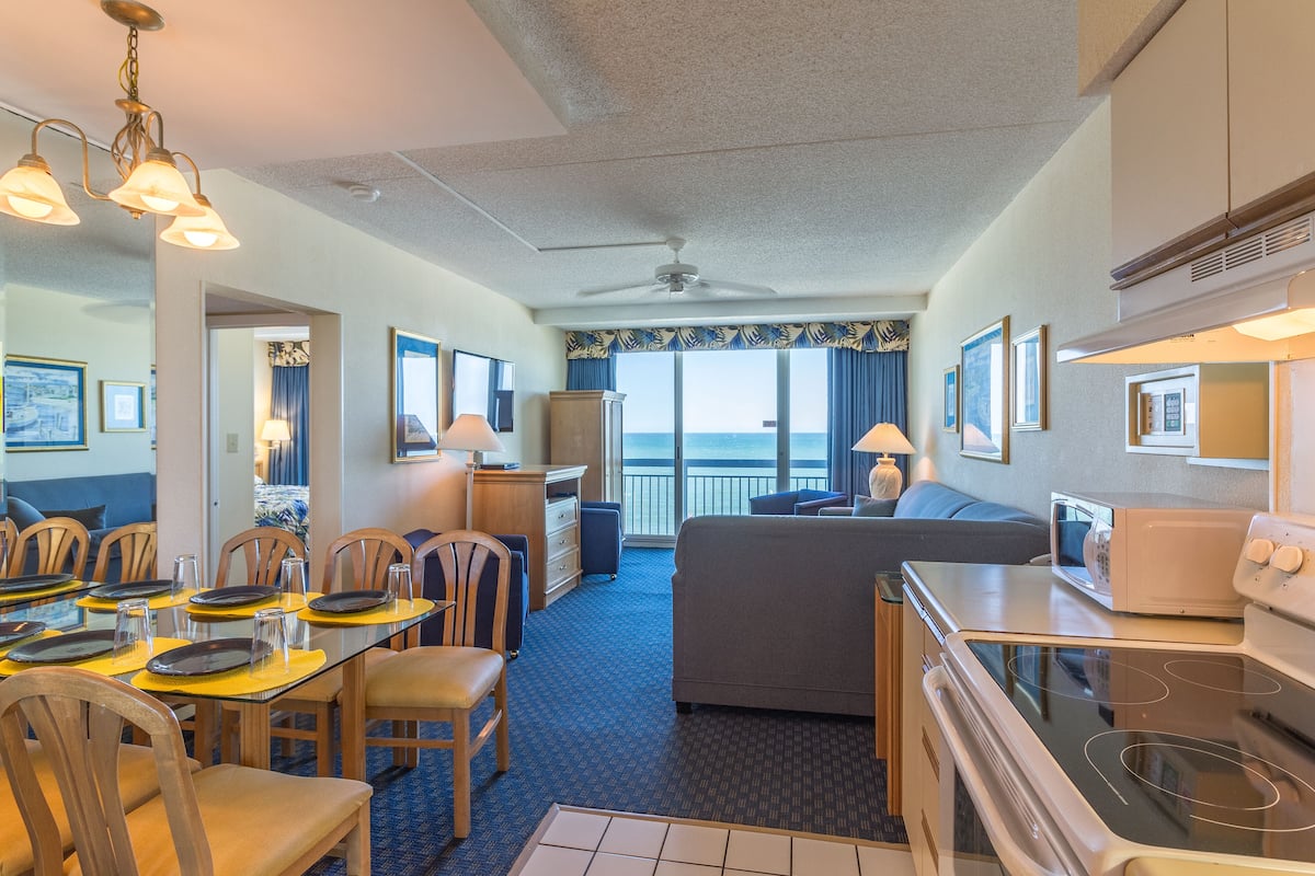 A hotel room with a dining area, a living area, a kitchenette, and a balcony with a view.