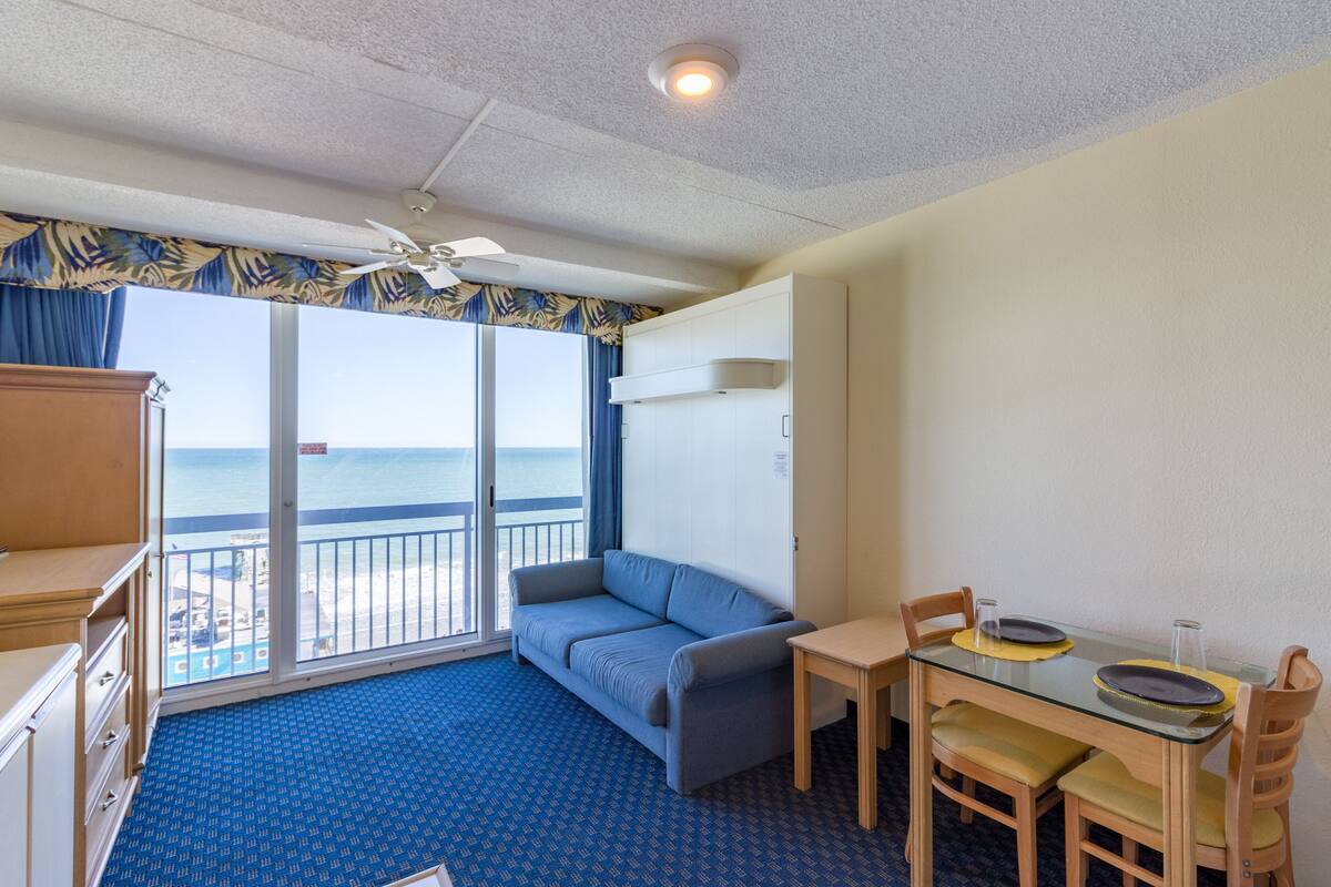 A room with a balcony, a sofa, a table with chairs, and a ceiling fan.