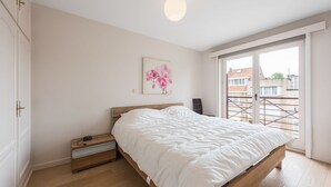 A bedroom with a wooden bed, a bedside table, a nightstand, a white wall, a pink orchid painting, and a view through a sliding glass door. - La Bohème 204 (Koksijde)