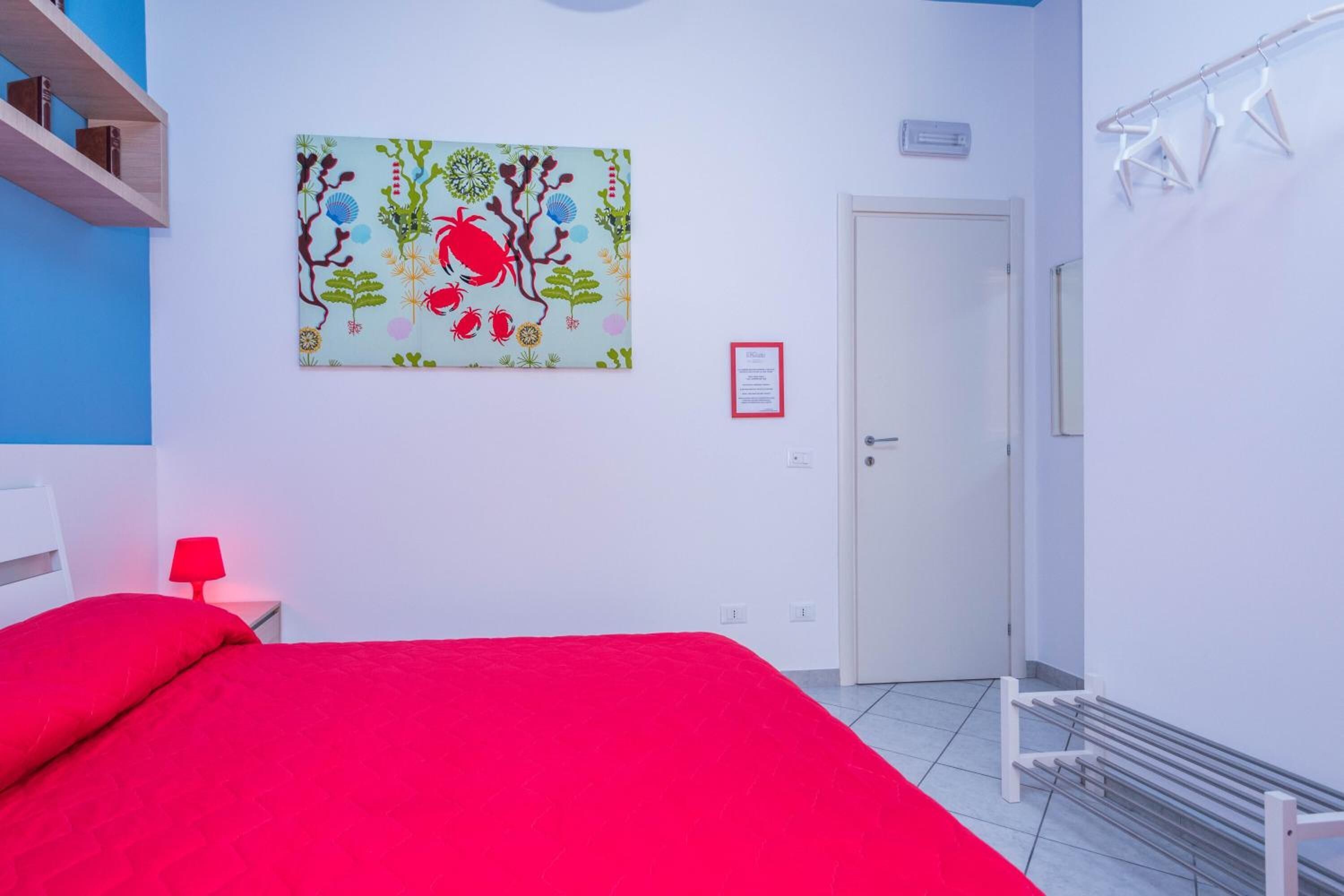 A bedroom with a red bed, a wall-mounted painting, a white door, and a laundry rack.