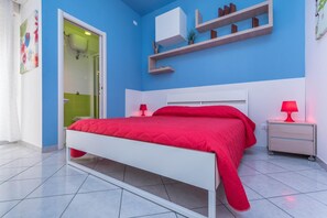 Une chambre avec un lit blanc, une literie rouge, une table de chevet avec une lampe et une étagère avec des livres et une plante.
