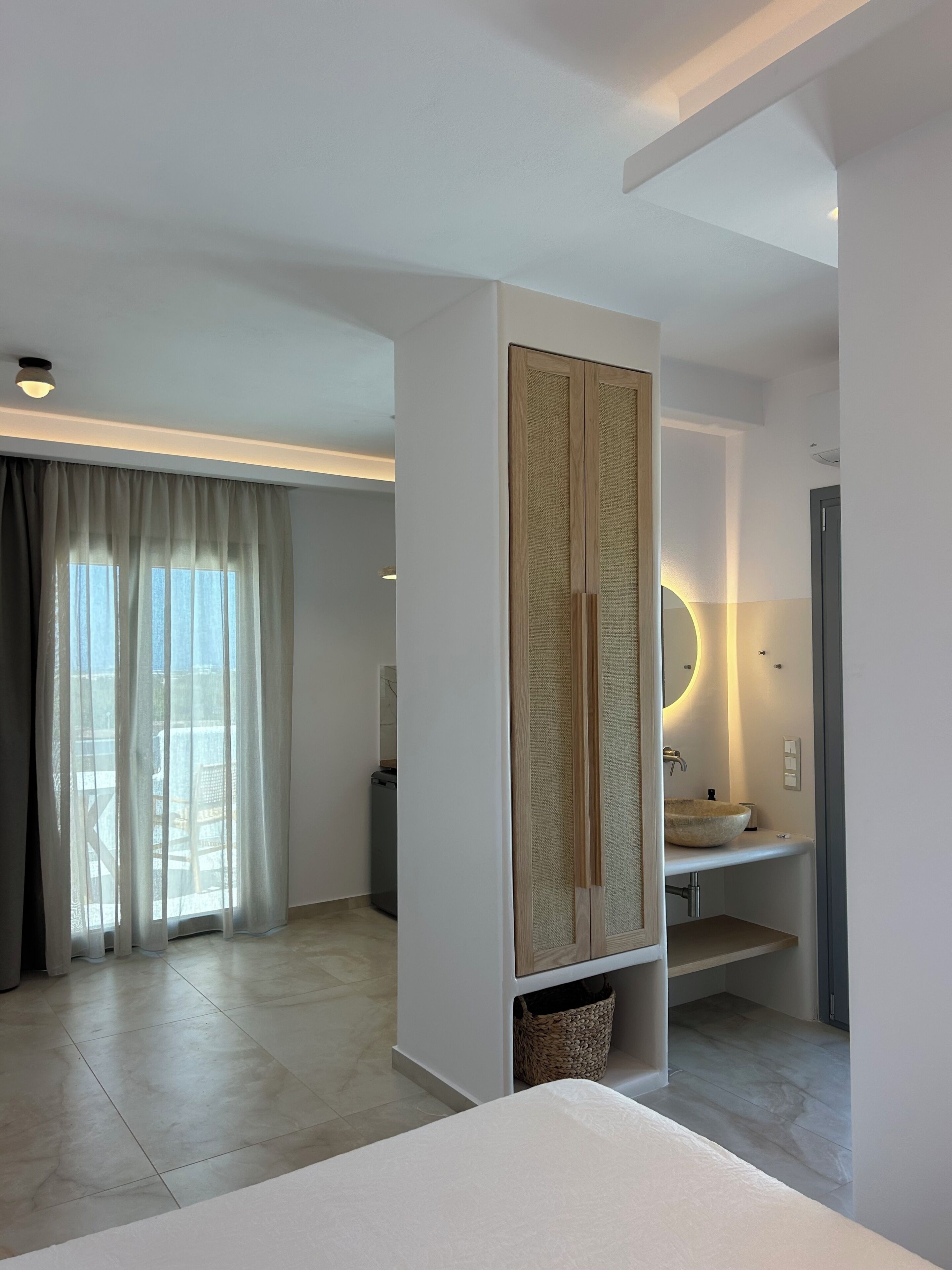 A modern hotel room with a large bed, a bathroom with a round mirror and sink, a wardrobe, and a view through a window with sheer curtains.