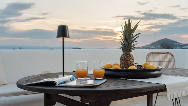 Una mesa redonda con una revista, dos vasos de jugo de naranja, una piña y un bol de fruta en un balcón con vistas a un paisaje montañoso.