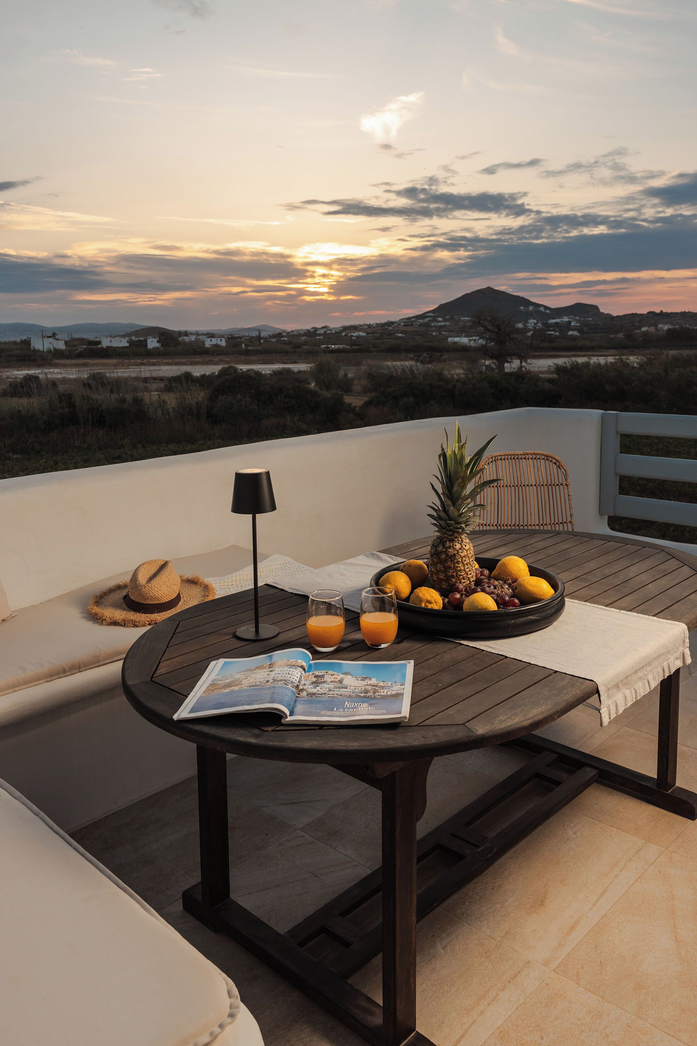 Une table ronde en bois, sur laquelle on trouve un magazine, un ananas, des citrons et un verre de jus d’orange, est placée sur un balcon offrant une vue sur une montagne et un paysage lointain.