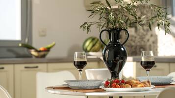 Una mesa de comedor con un florero de hojas verdes, dos copas de vino tinto, un plato de comida y un bol de fruta.