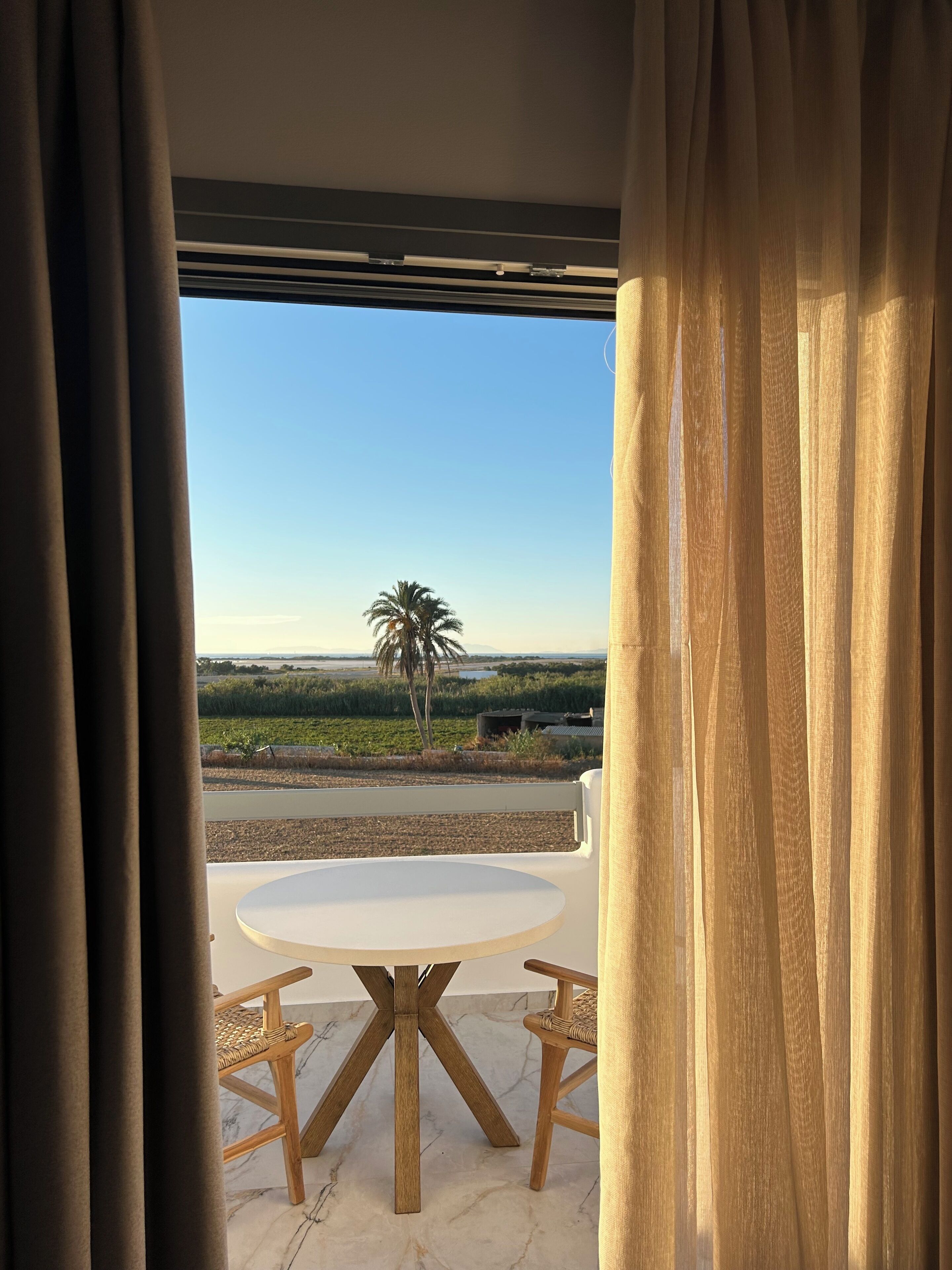 A view through a window with curtains, featuring a round table with chairs, a palm tree, and a distant landscape.