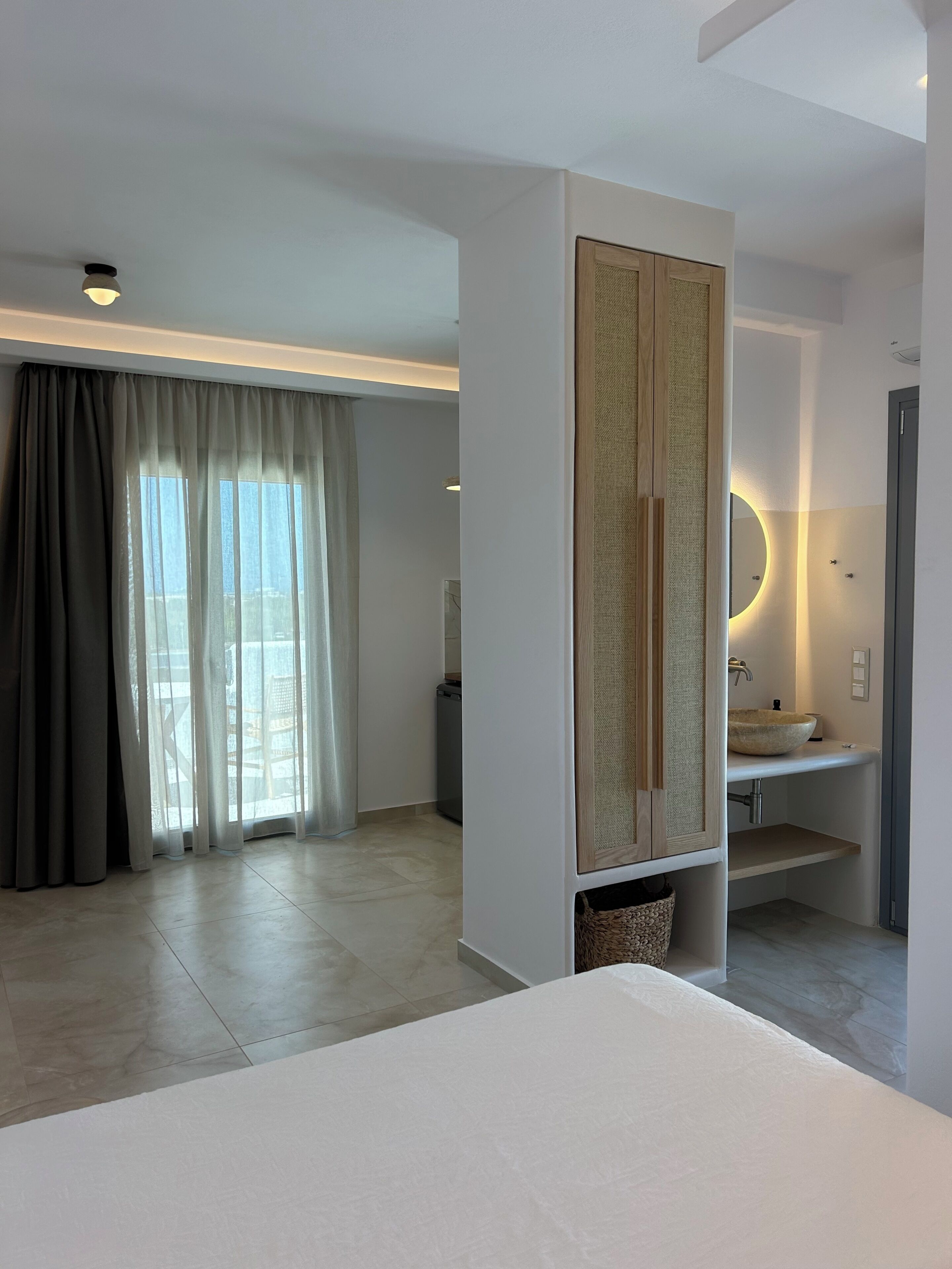 Ein modernes Hotelzimmer mit einem großen Bett, einer integrierten Garderobe, einem Badezimmer mit rundem Spiegel und Waschbecken sowie Blick durch transparente Vorhänge.
