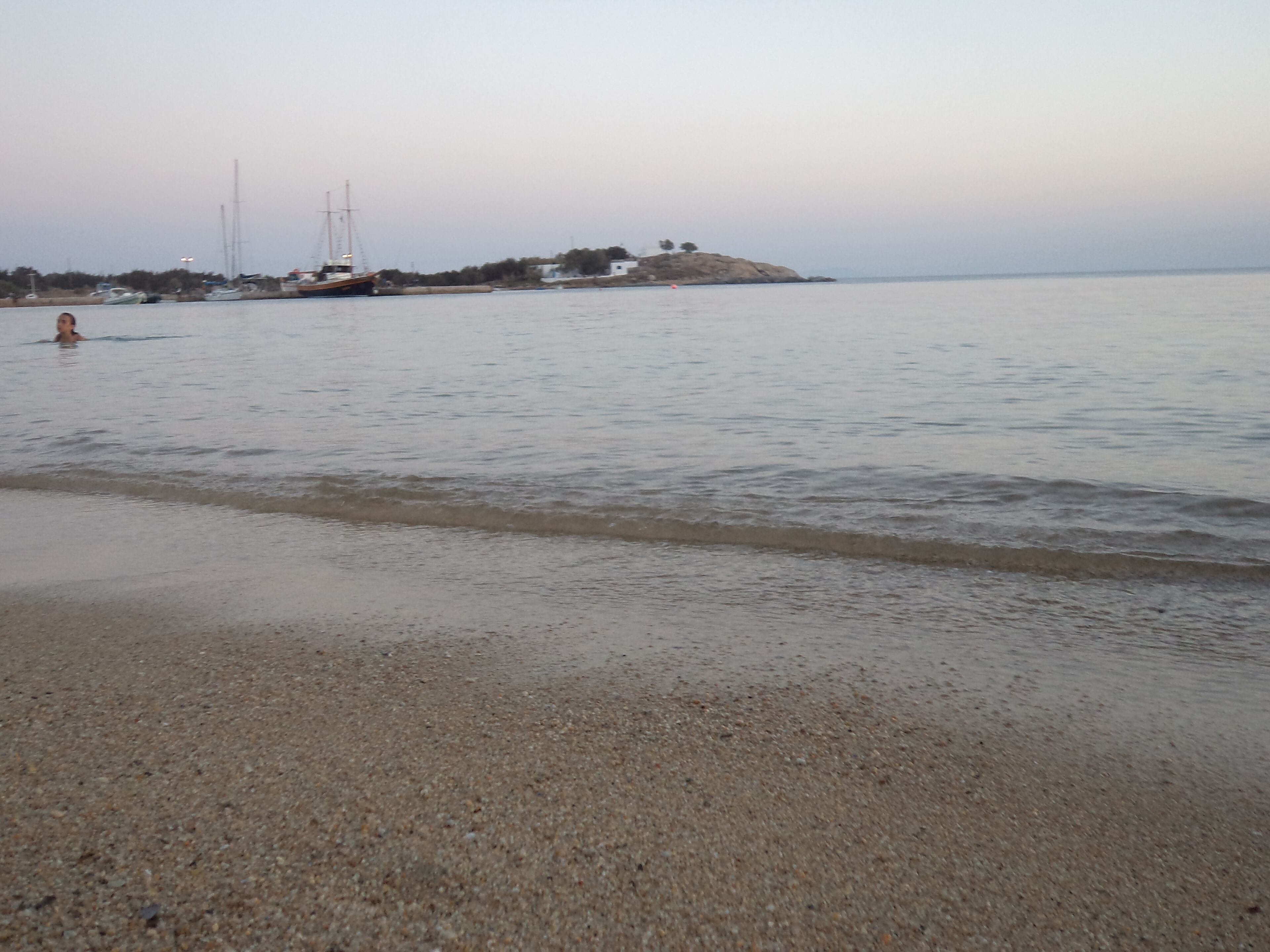 A beach with calm water, pebbles, and a distant view of boats and buildings.
