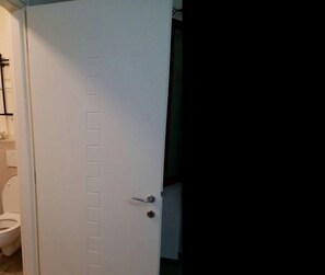 Puerta blanca con manija plateada y cerradura, que da acceso a un baño con inodoro y zona de ducha.