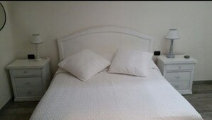 Een net opgemaakt bed met witte lakens en kussens, met aan weerszijden nachtkastjes met lampen en een klein lade.