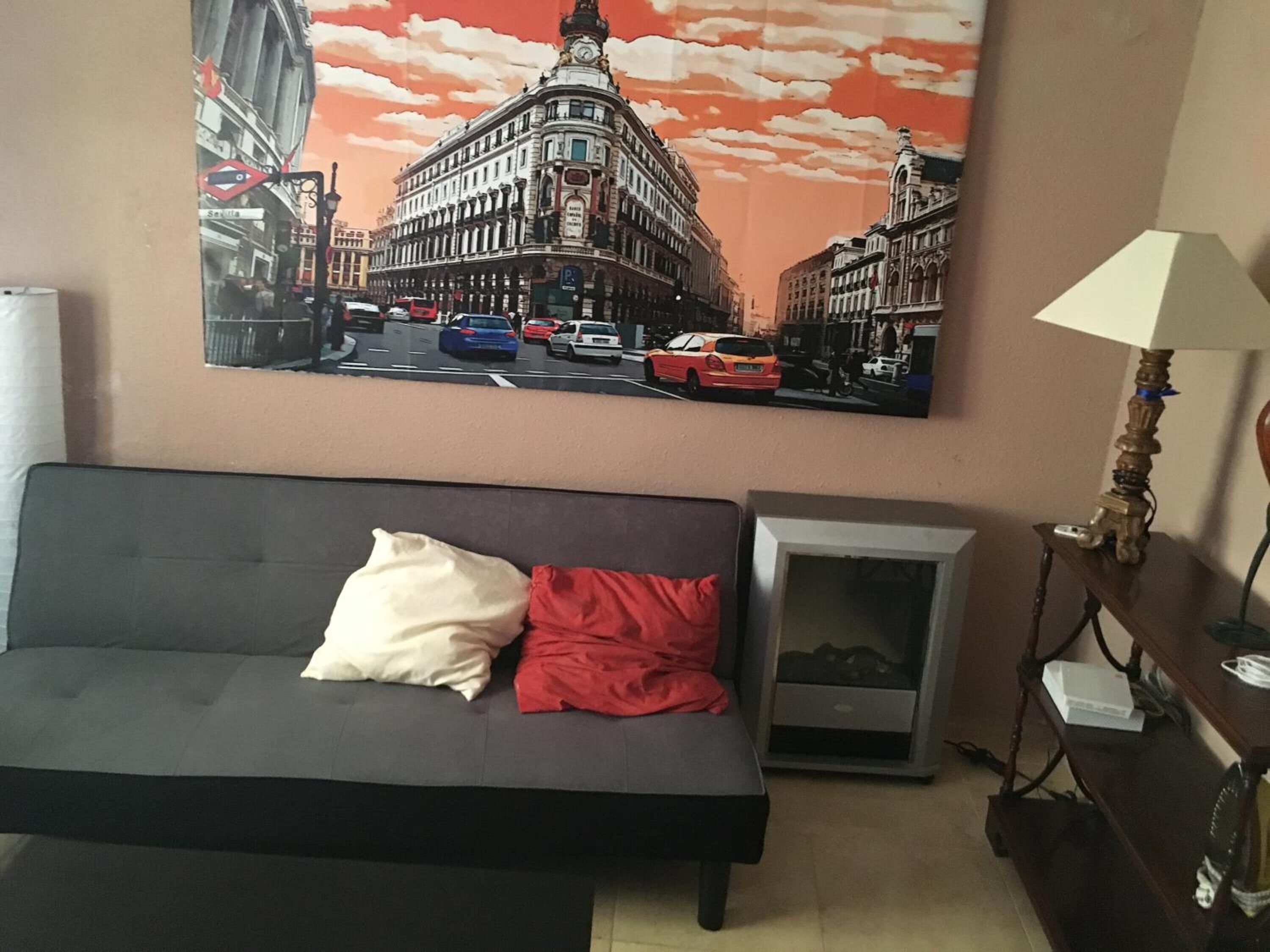 A living room with a black sofa, a red pillow, a lamp, and a painting of a cityscape.