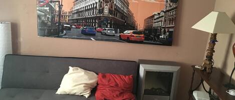 A living room with a black sofa, a red pillow, a lamp, and a painting of a cityscape.