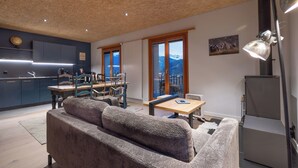 A modern living room with a sofa, dining table, and chairs, a kitchen with blue cabinets, and a large window with a mountain view. - B&B La Forge de Diogne (Crans-Montana)