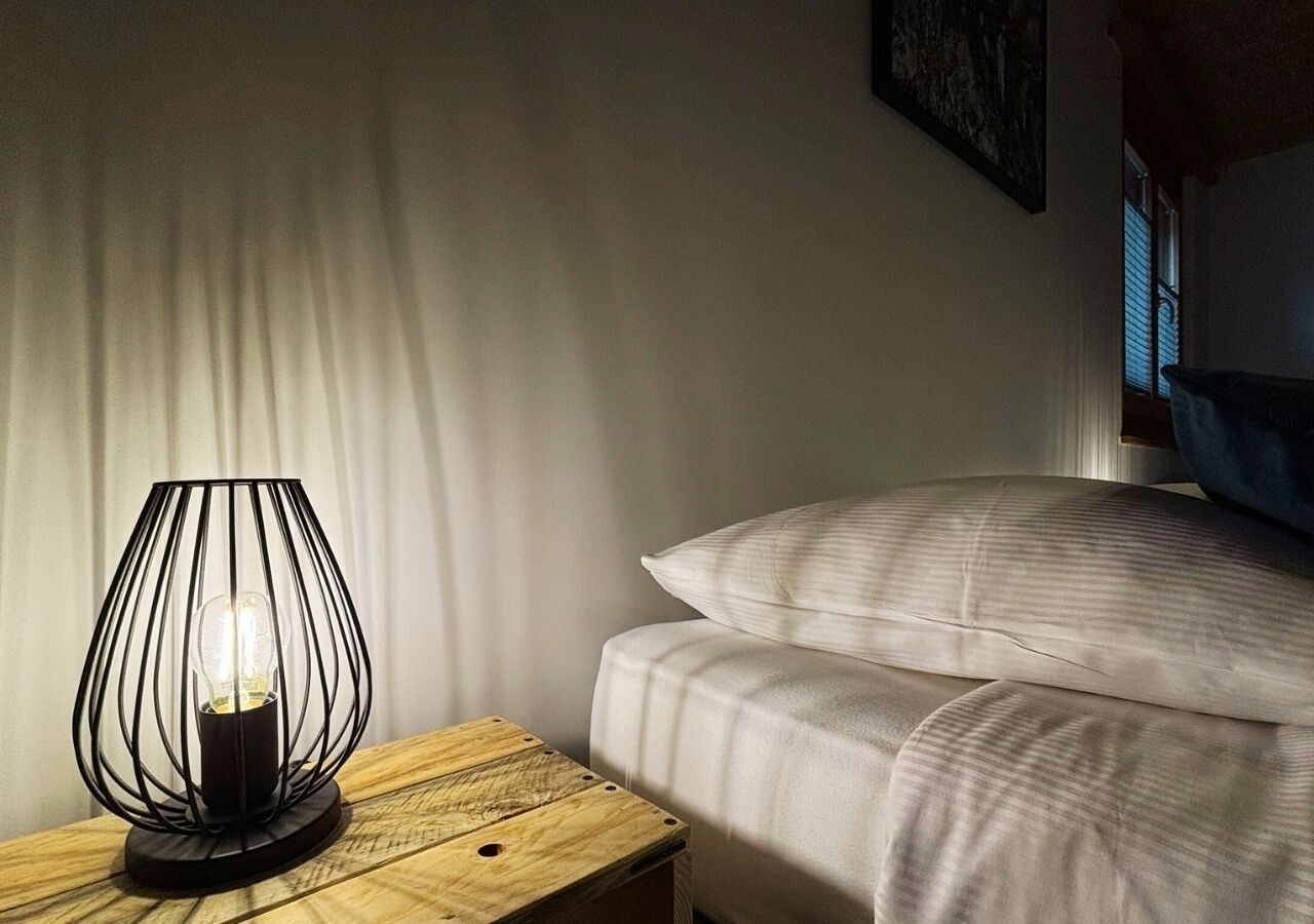 Un letto con biancheria bianca, un comodino con una lampada e una finestra con persiane.