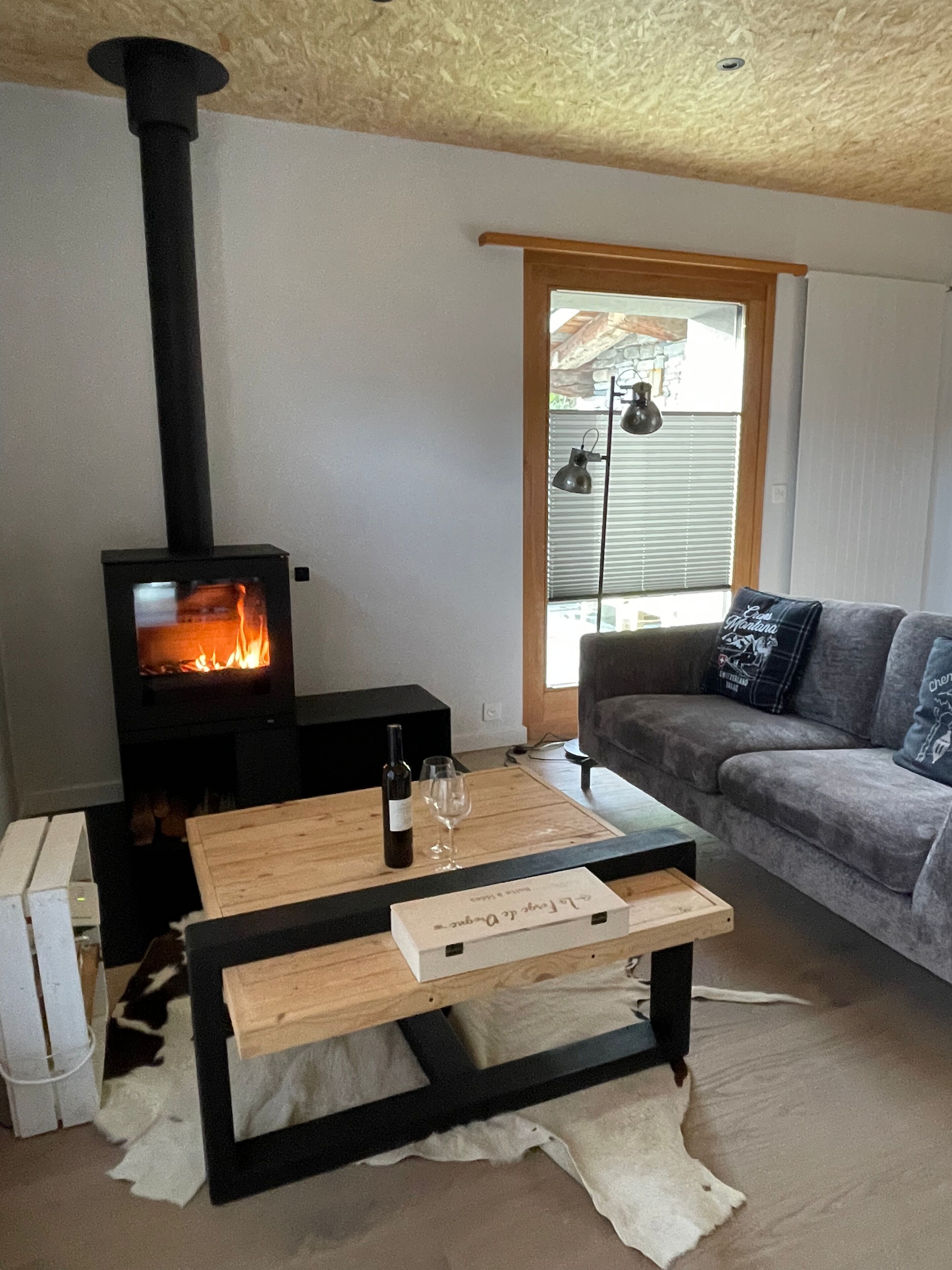 A cozy living room with a wood-burning stove, a sofa, a coffee table, and a window with blinds.