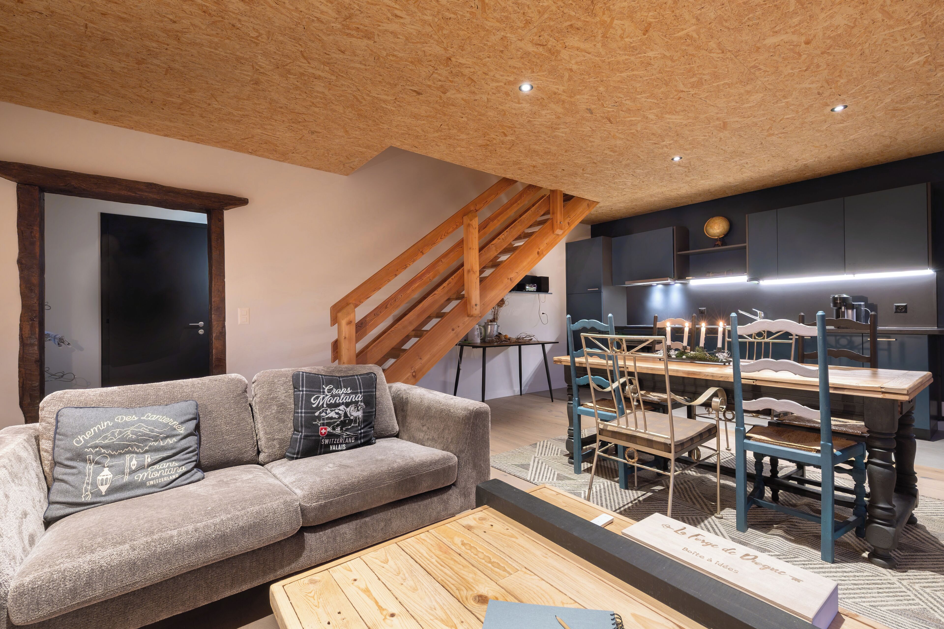 Een moderne woonkamer met een houten plafond, een grijze bank met decoratieve kussens, een houten salontafel, en een eethoek met een blauwe stoel en een tafellamp.