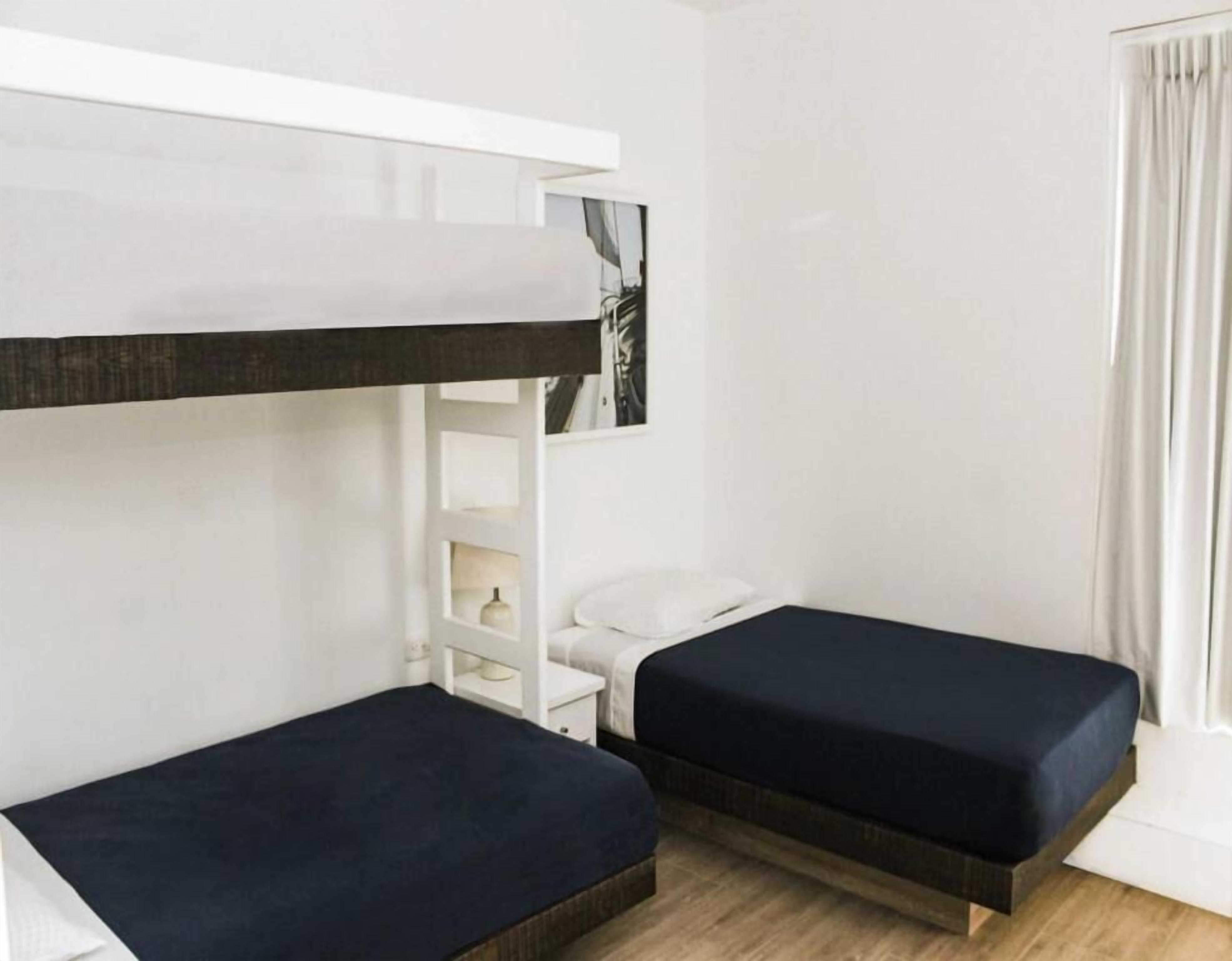 Una habitación con dos camas individuales, una mesita de noche blanca, un estante blanco y una ventana con cortinas.