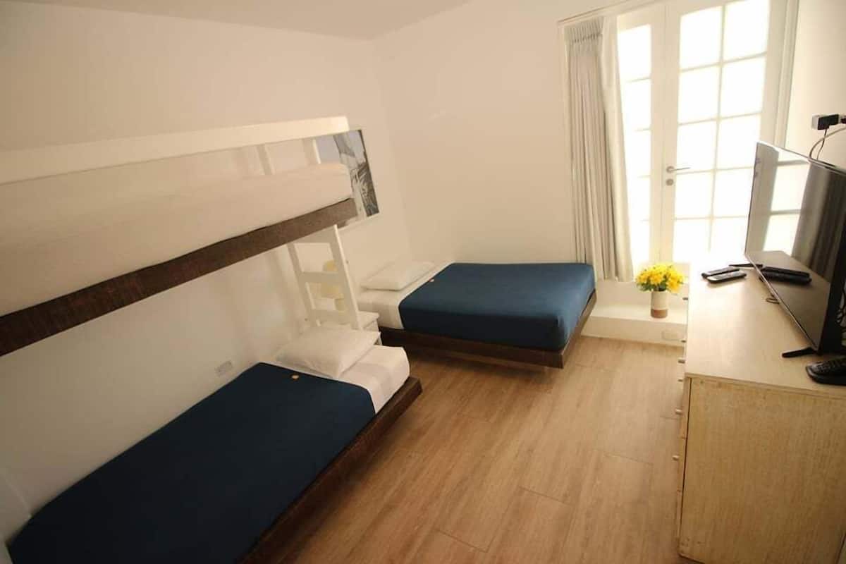 Ein kleiner, gut beleuchteter Raum mit zwei Einzelbetten, einem Schreibtisch mit Fernseher und einem Fenster mit Vorhängen.
