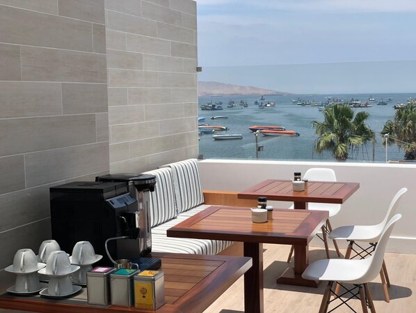 Un balcón con zona de estar, cafetera y vista a barcos en el agua.