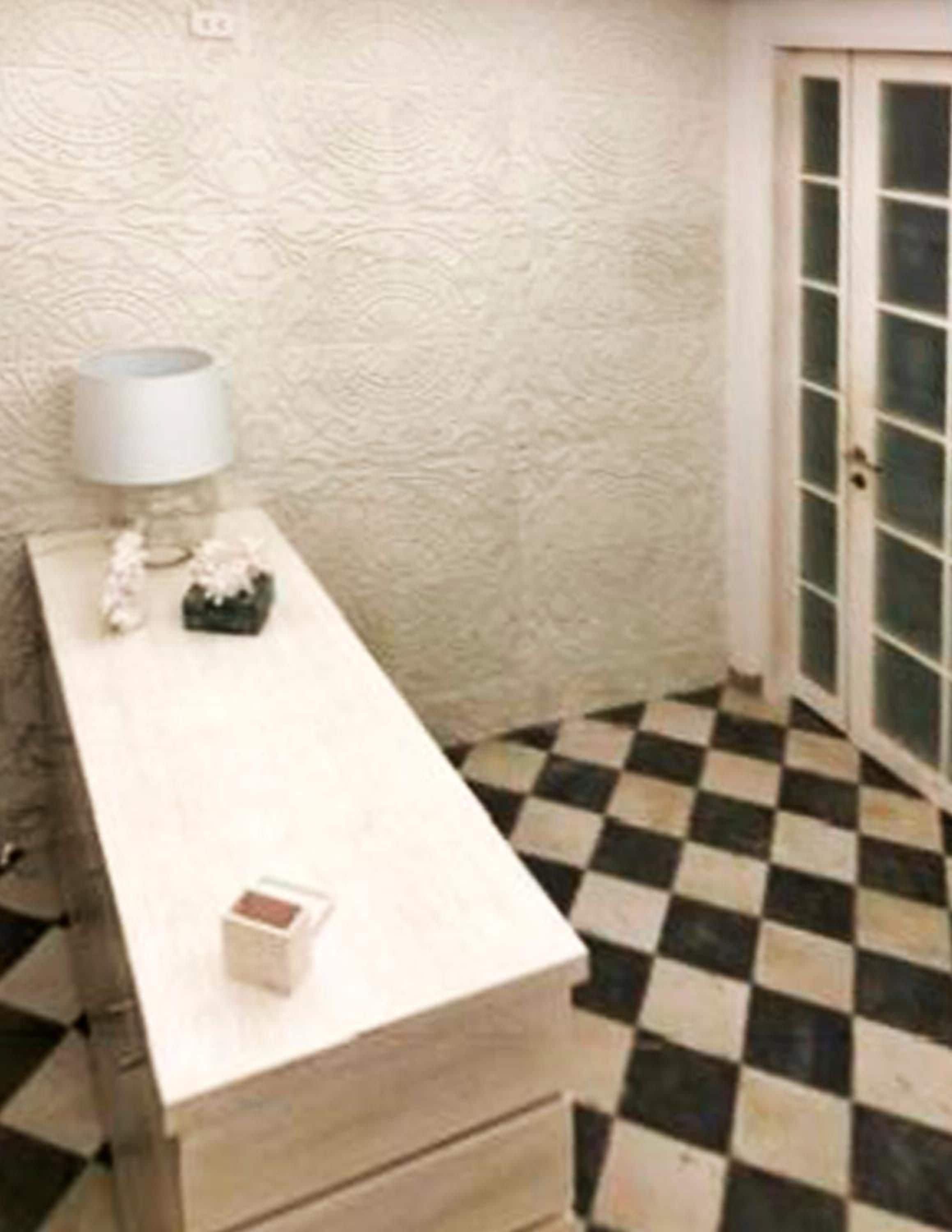 A room with a checkered floor, a white textured wall, a small table with a lamp, and a door with glass panels.