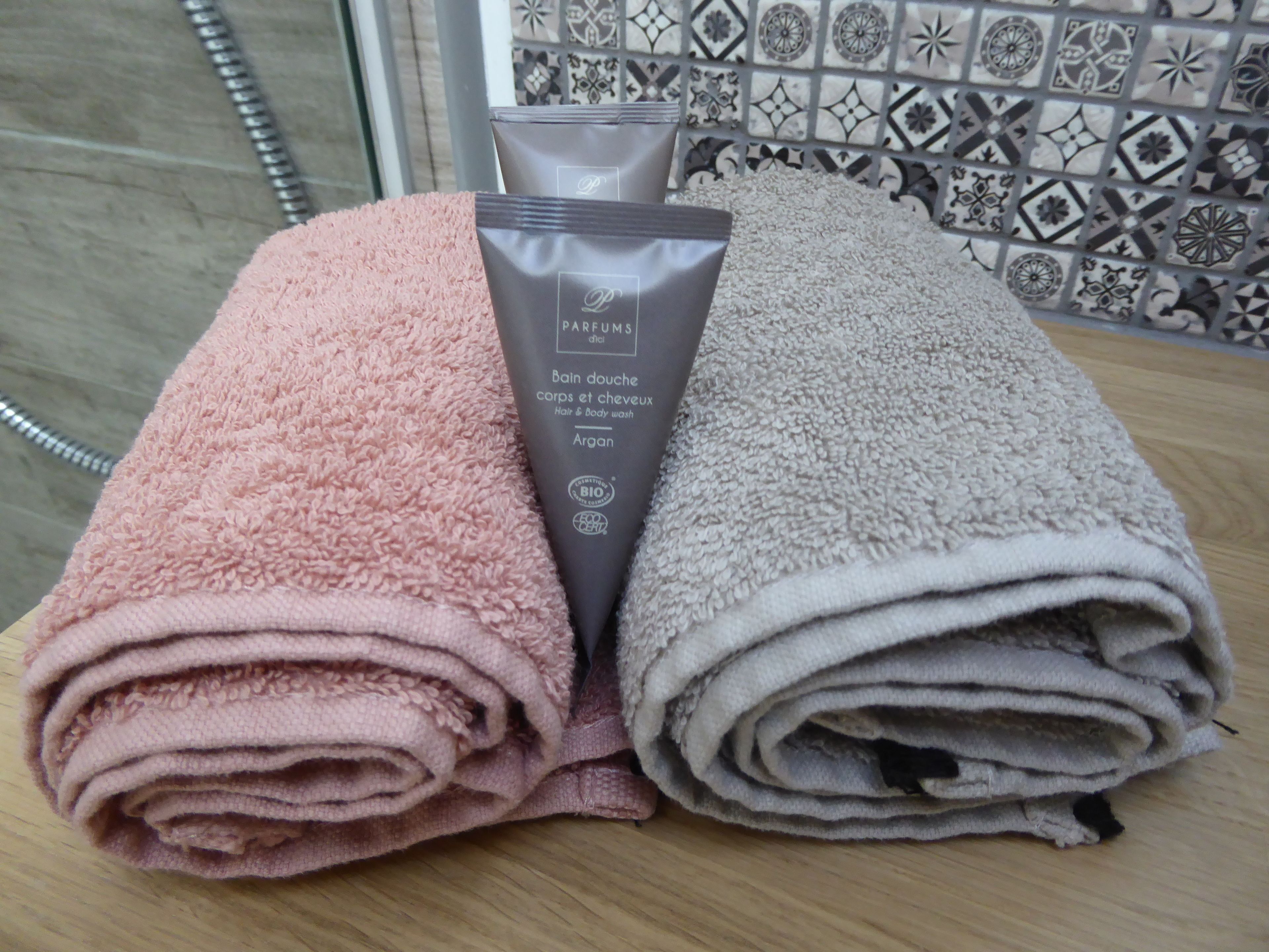 Dos toallas enrolladas, una rosa y otra color beige, sobre una superficie de madera, con un fondo de baño.