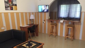 A living room with a sofa, coffee table, TV, and a kitchen area with stools and a counter. - Hans Cottage Botel (Cape Coast)