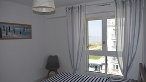 Ein Schlafzimmer mit Bett, Stuhl, einem Fenster mit Vorhängen, einem Gemälde an der Wand und Blick auf Gebäude und Grünflächen.