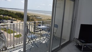 Ein Balkon mit Glastür, Tisch und zwei Stühlen, mit Blick auf einen Strand und eine Straße mit Autos.