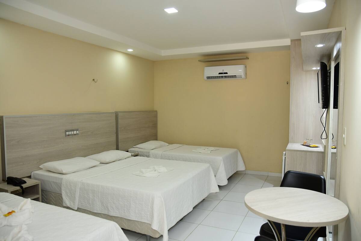 A hotel room with two beds, a small table, a desk with a chair, and an air conditioning unit on the wall.