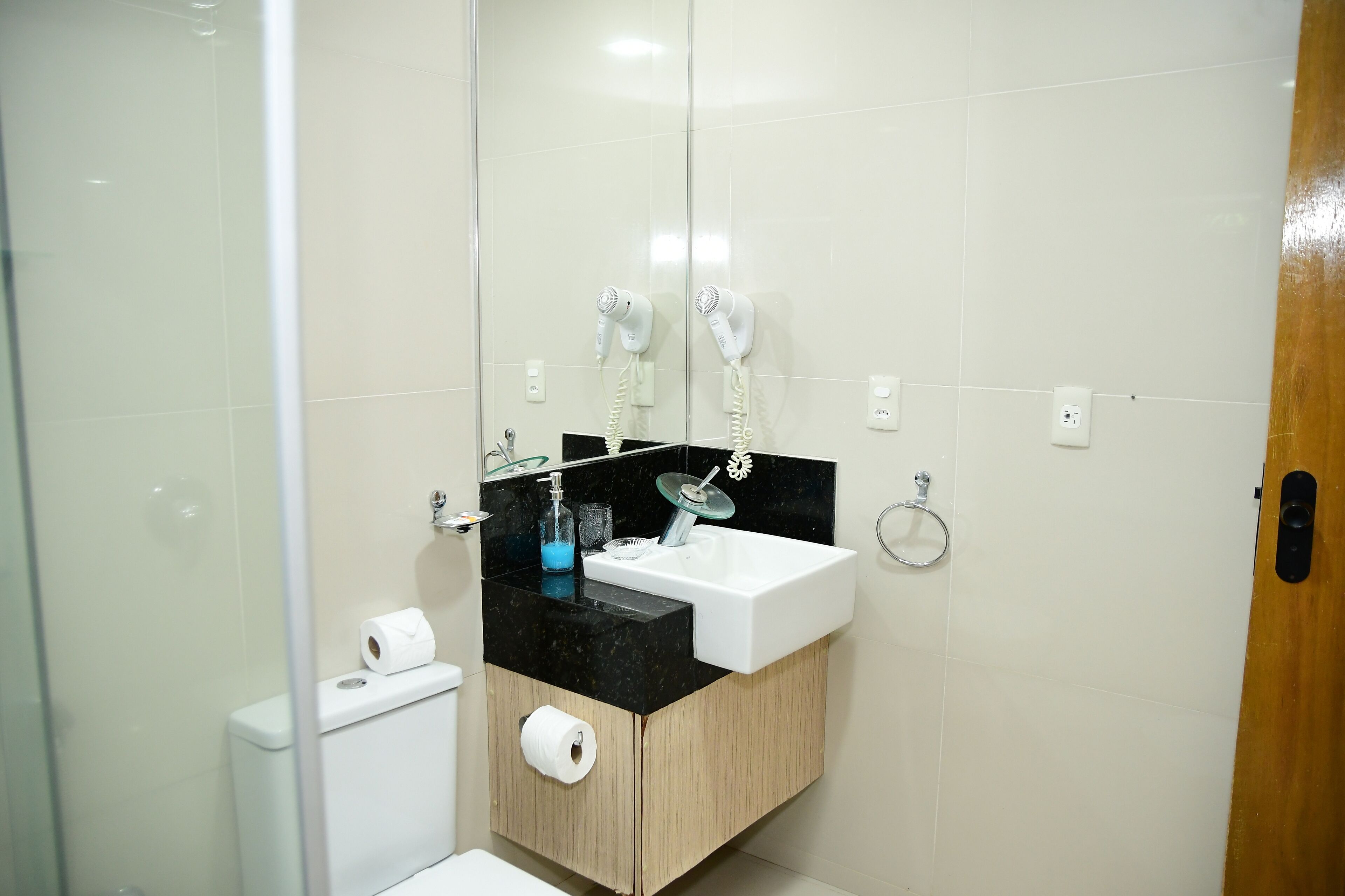 A bathroom with a white sink, black countertop, and a mirror above it. There is a toilet with a roll of toilet paper and a glass shower enclosure.