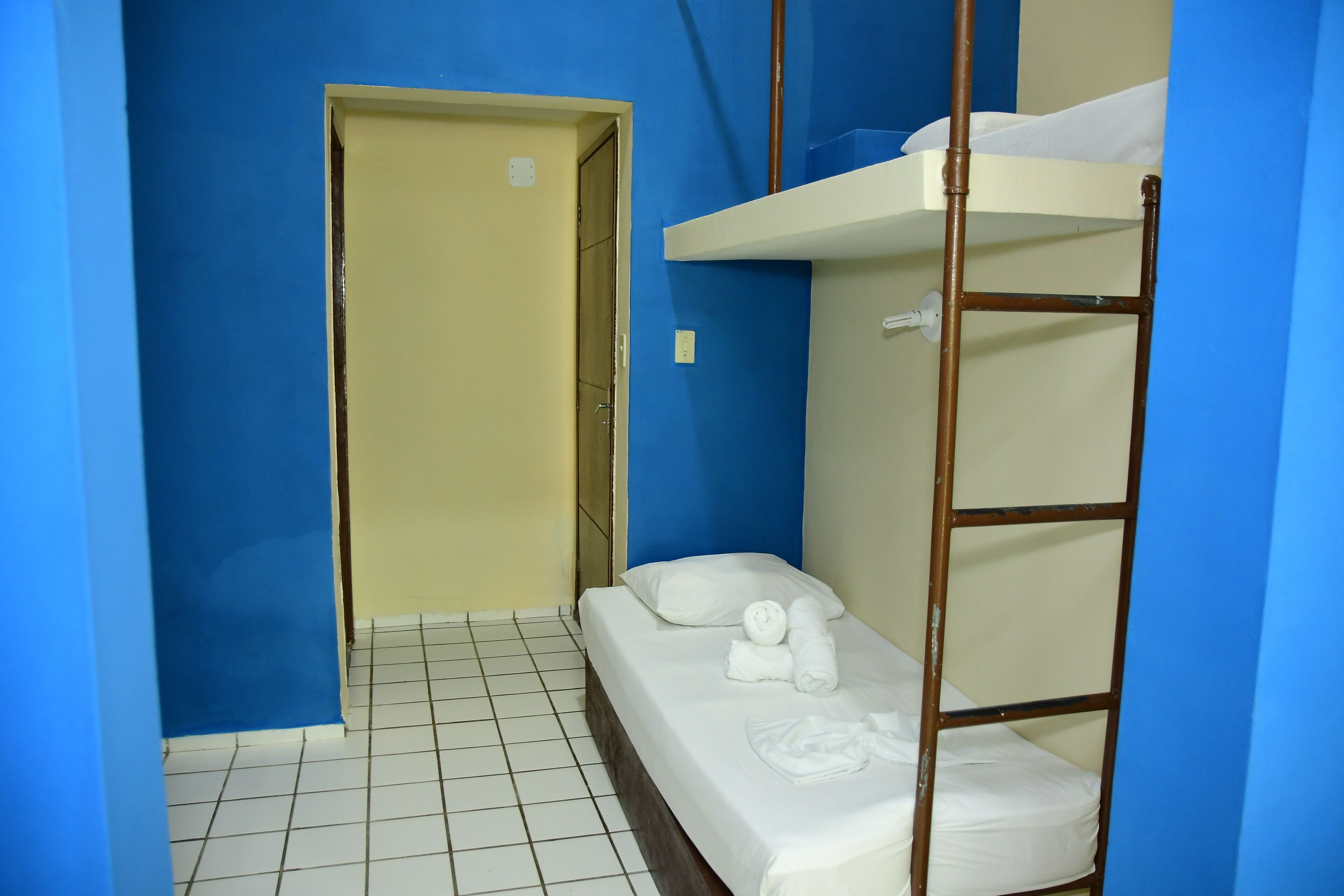Una habitación pequeña y funcional con una litera y una cama individual. Hay una puerta que da a otra habitación y el suelo está cubierto de baldosas.
